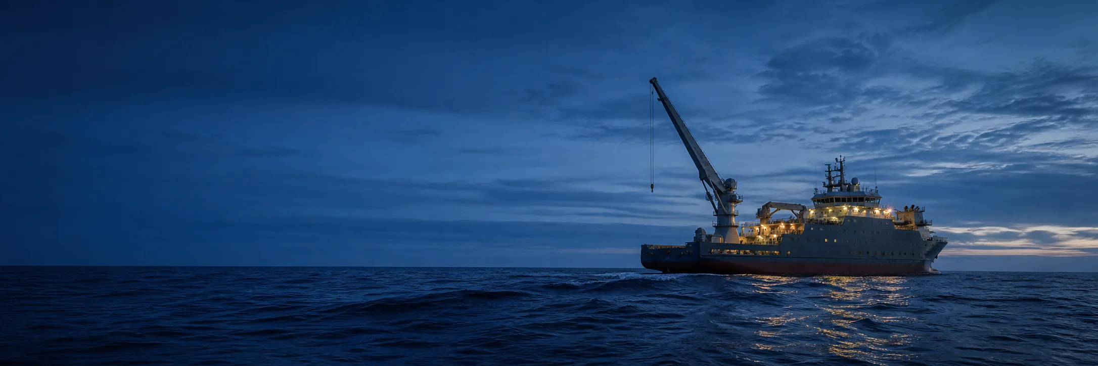 An offshore construction vessel at blue hour working in a moderate beam sea, its crane boom raised against the sky