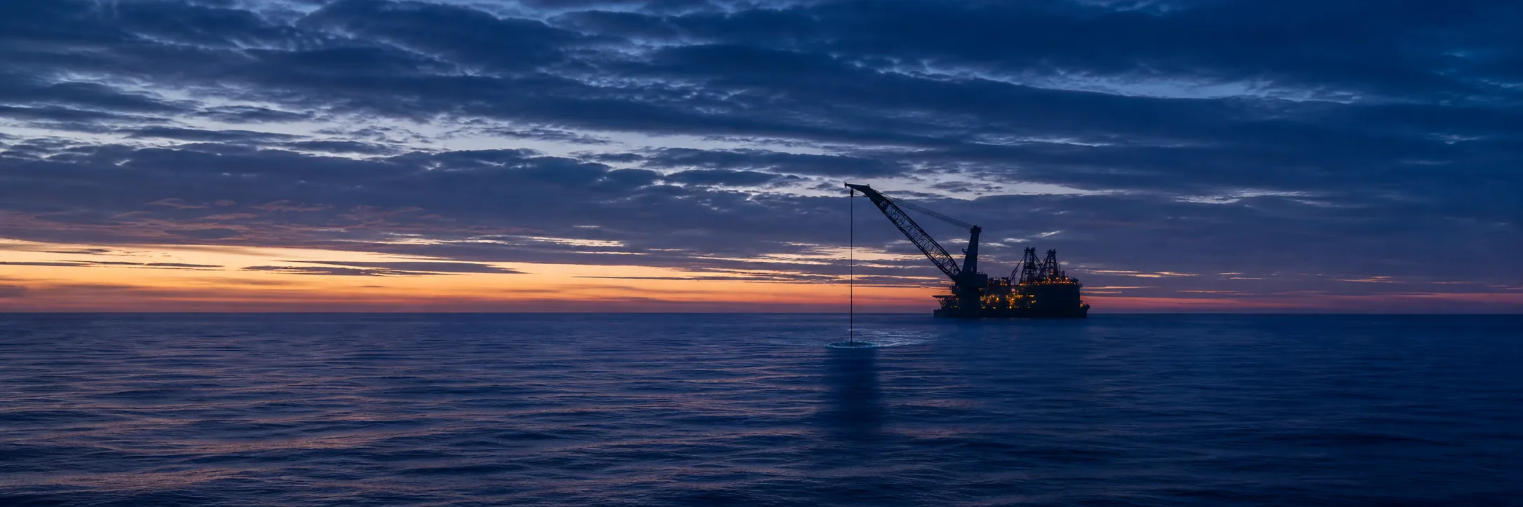 Offshore tungløftfartøy i blåtimen som senker en undervannskonstruksjon i én stram kranwire gjennom havoverflaten