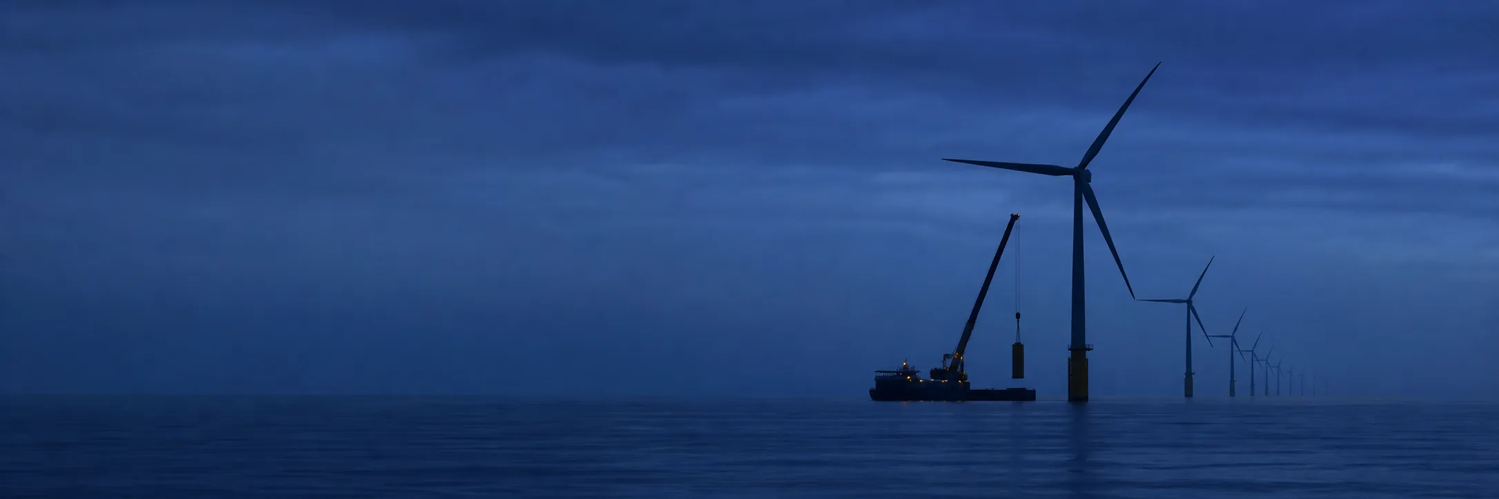 Una fila de aerogeneradores marinos se pierde en la neblina de la hora azul, con un buque de instalación junto a la cimentación más cercana
